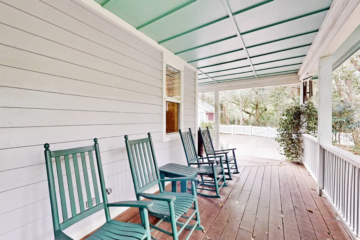 This image displays a cozy covered porch with classic wooden rocking chairs, a wooden floor, and a charming green ceiling, surrounded by lush trees. Natural light filters through, and the space feels serene and welcoming—inviting guests to relax, read, or socialize while immersed in nature.