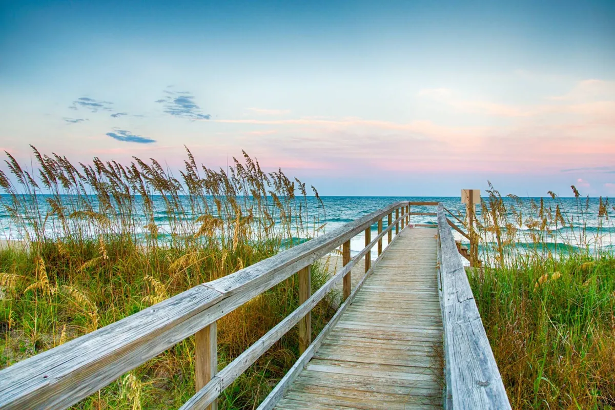 A serene boardwalk leading to the ocean, bordered by dune grass and framed by a soft, cloud-filled sky. The scene invites leisurely walks and moments of reflection by the shore, reinforcing the island’s relaxed coastal vibe.