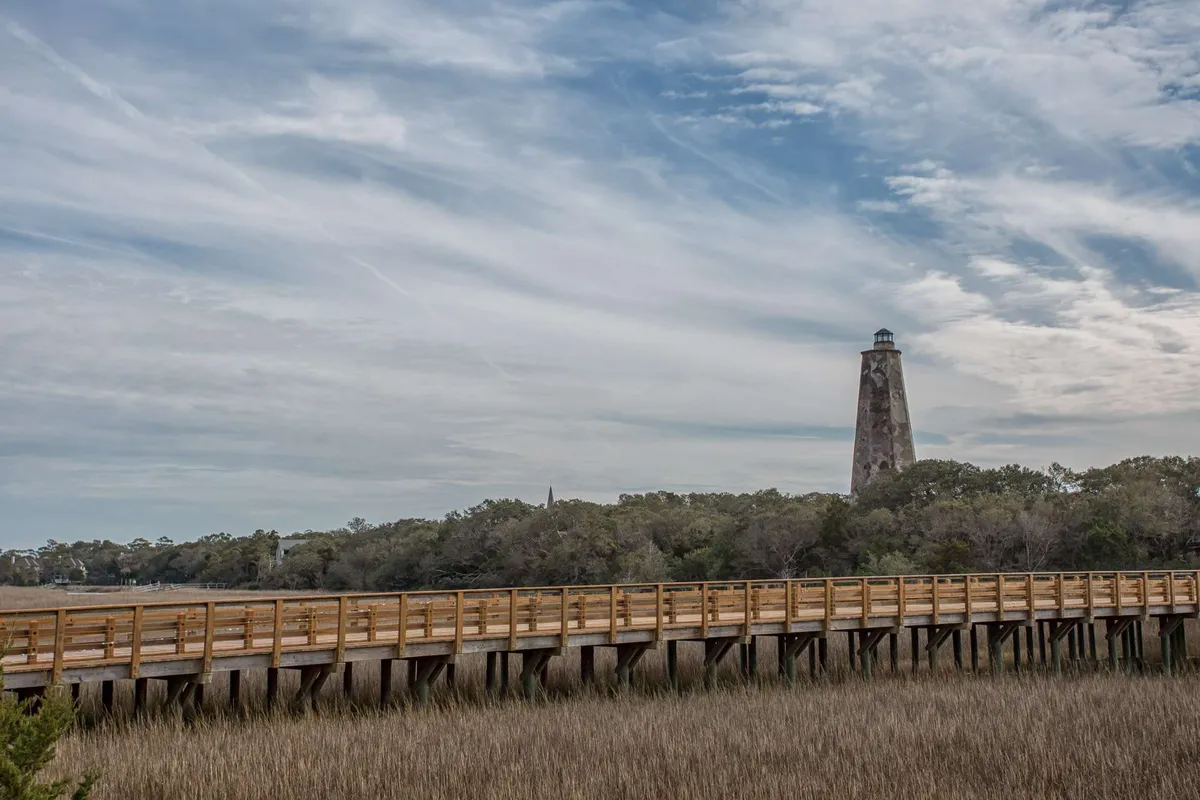An exterior view featuring the iconic island lighthouse, a wooden boardwalk winding through marshland, and a dramatic cloudy sky above. The natural surroundings create a tranquil and inviting environment for outdoor exploration and discovery.