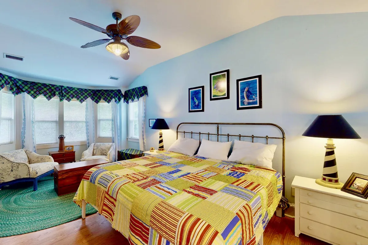 A comfortable bedroom featuring a colorful quilt on the bed, large windows that flood the room with natural light, and tasteful artwork on the walls. The room exudes warmth and tranquility, ideal for restful sleep or quiet relaxation.