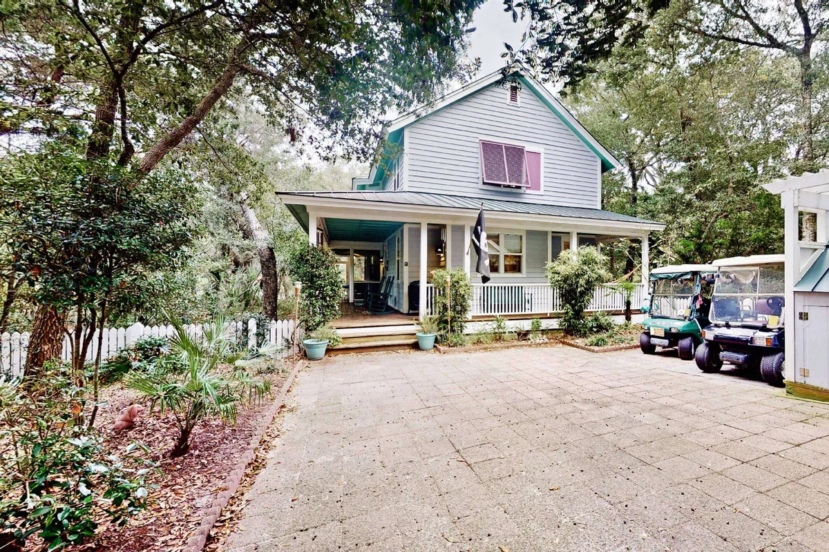 A charming coastal cottage with a spacious screened front porch surrounded by lush greenery, featuring two golf carts parked nearby for easy island exploration. The inviting porch is furnished for relaxation, perfect for enjoying morning coffee or unwinding after a day outdoors.