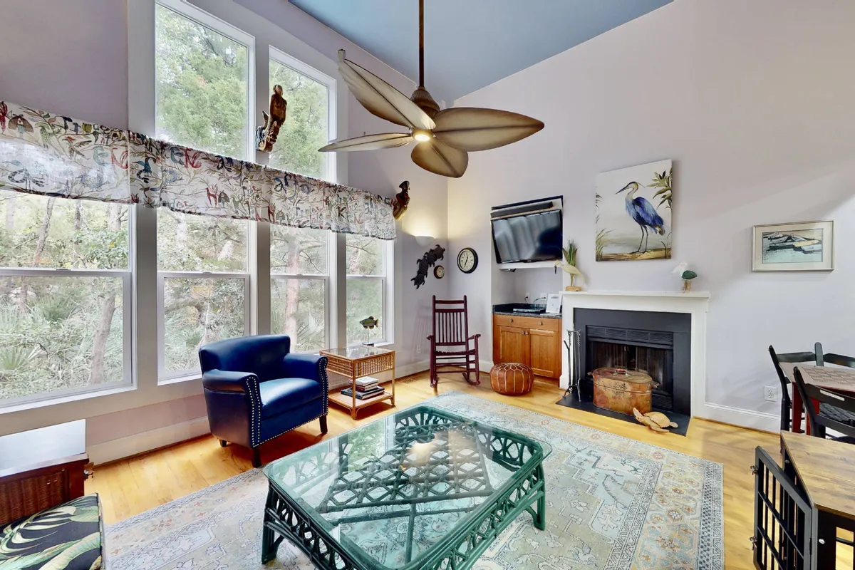 This image features a comfortable living room with large windows that flood the space with natural light. The room is highlighted by a blue ceiling, a decorative ceiling fan, a central coffee table, and thoughtfully selected artwork on the wall. The seating is arranged to encourage relaxation or conversation, and the overall mood is serene and inviting.