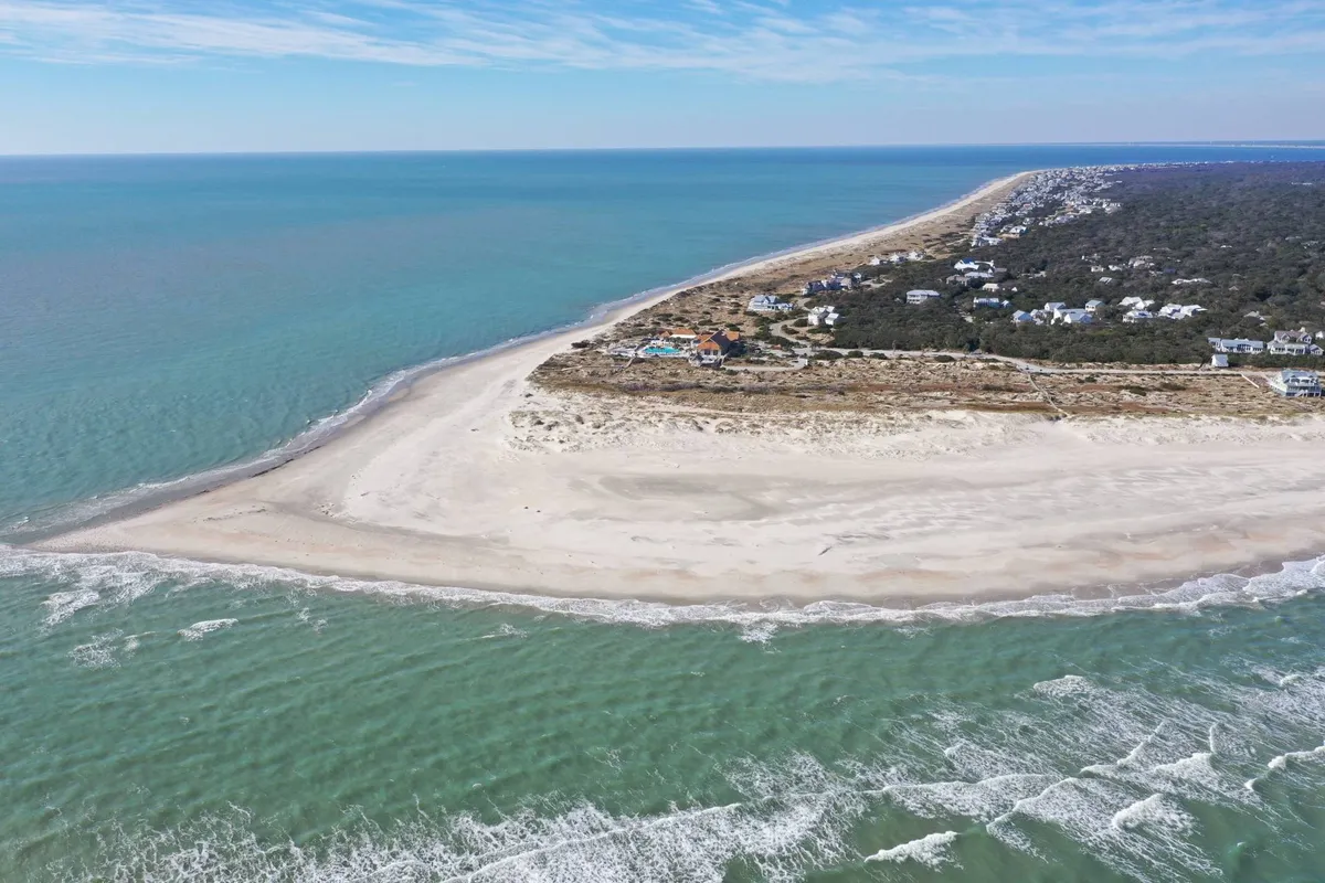 A tranquil stretch of sandy beach meeting gentle ocean waves, bordered by coastal vegetation and dotted with distant seaside houses. The scene invites activities like swimming, collecting seashells, and enjoying picnics on the sand. The mood is serene and family-friendly, perfect for a fun beach day.