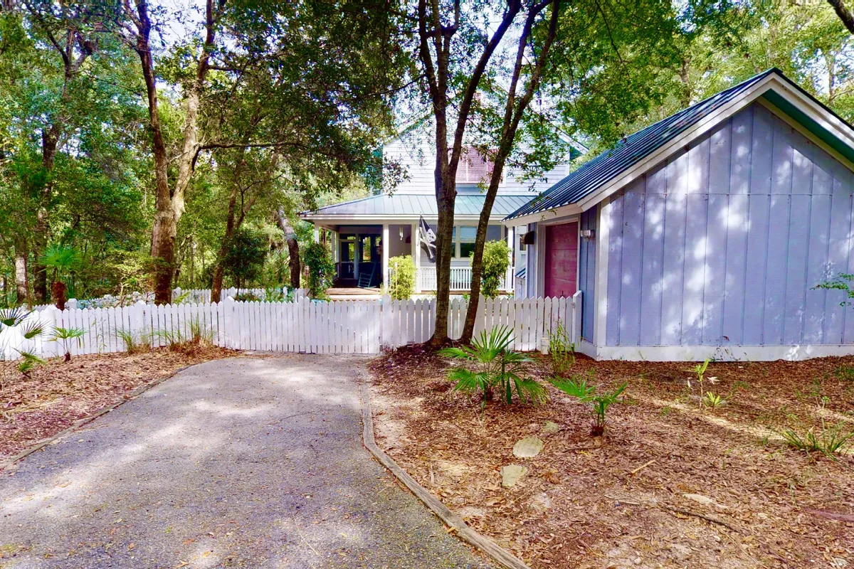 A charming exterior view of Dundee's Den, featuring a classic white picket fence that fully encloses the lush front yard. The inviting porch sits beneath a shady tree canopy, offering a perfect spot to relax and keep an eye on your playful pup. The setting conveys a welcoming, secure environment ideal for families with pets.