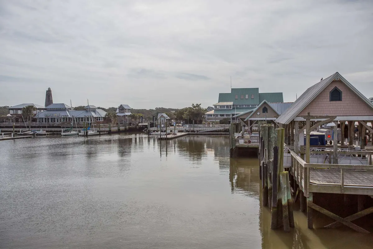 A tranquil waterfront scene featuring docks and boathouses, with the iconic lighthouse visible in the distance. The calm water reflects the sky, creating a serene atmosphere ideal for leisure walks, boating, and waterfront dining.
