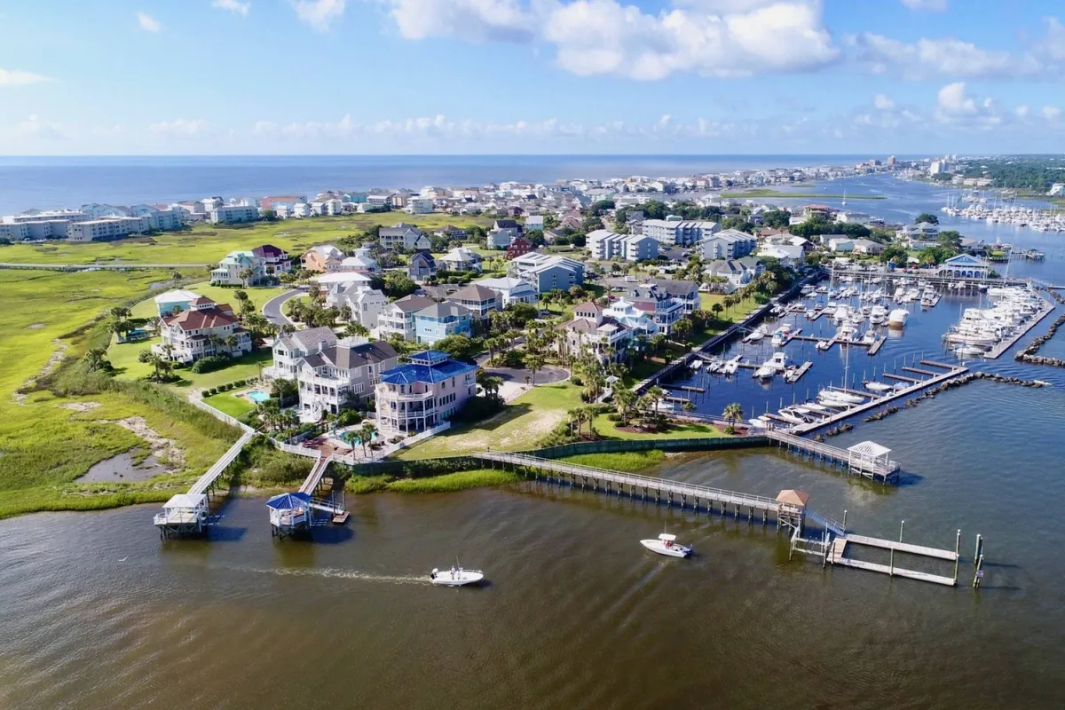A picturesque marina scene featuring boats docked along the water, lush green spaces, and charming coastal homes in the background. The vibrant greenery and waterfront setting evoke the coastal lifestyle and local market culture of Bald Head Island.
