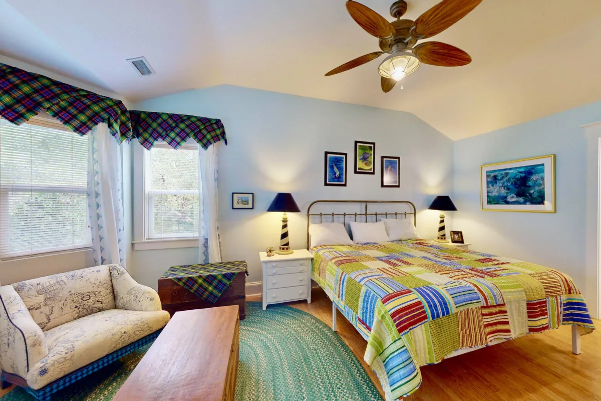 A cheerful bedroom with a colorful quilt, eye-catching artwork, a modern ceiling fan, and a comfortable armchair. The room is bright, welcoming, and offers a relaxing space for unwinding after a busy day.